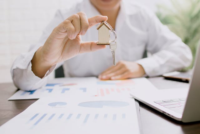 person holding a key with a house keychain while sat at a desk that has charts on it