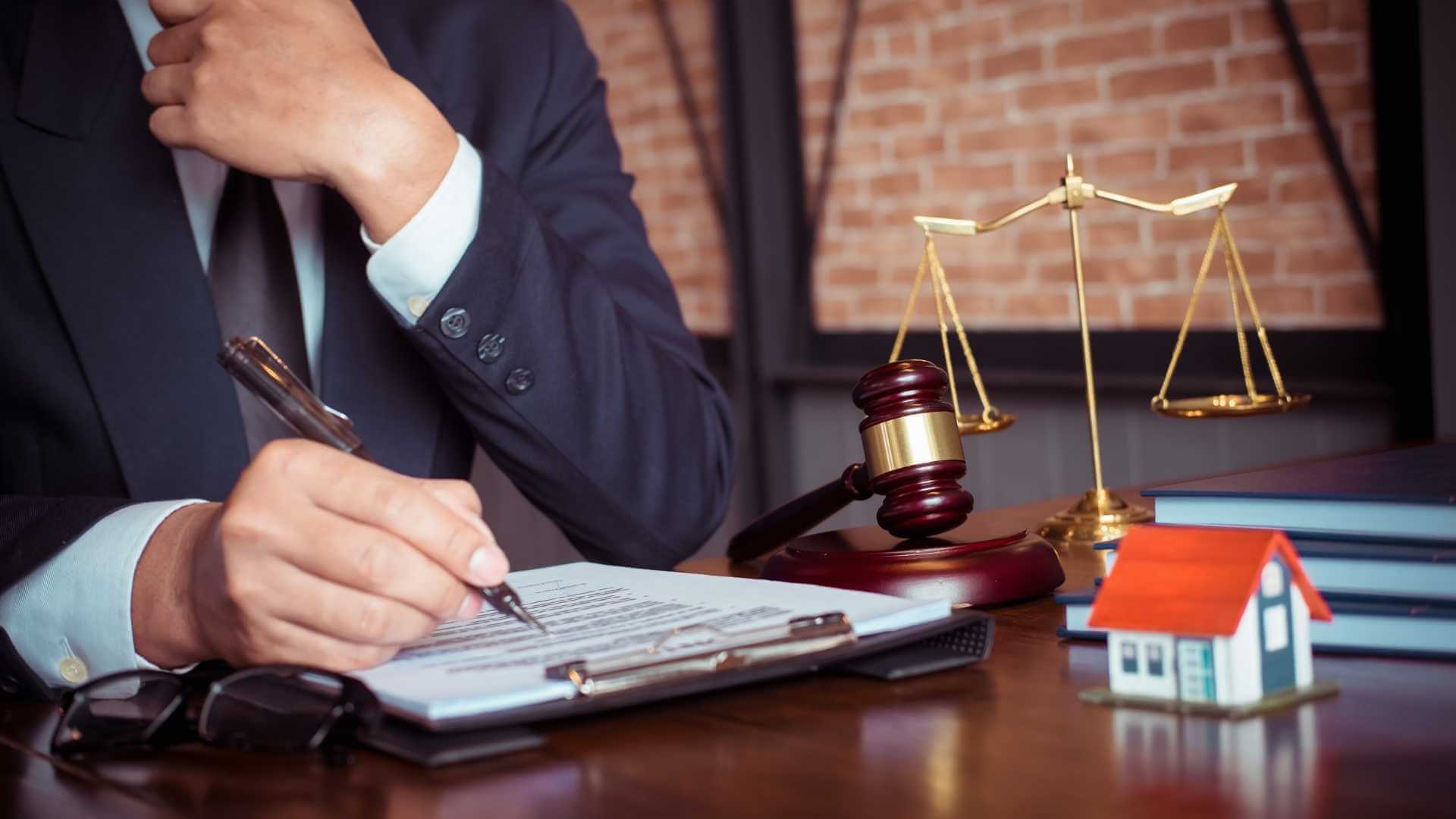 person signing legal document with gavel, balance scale, and house model on the desk